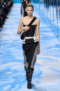 Model walking a blue-toned runway in a black strapless top and black‑and‑white checkered skirt, carrying a black crossbody bag with a wide strap.