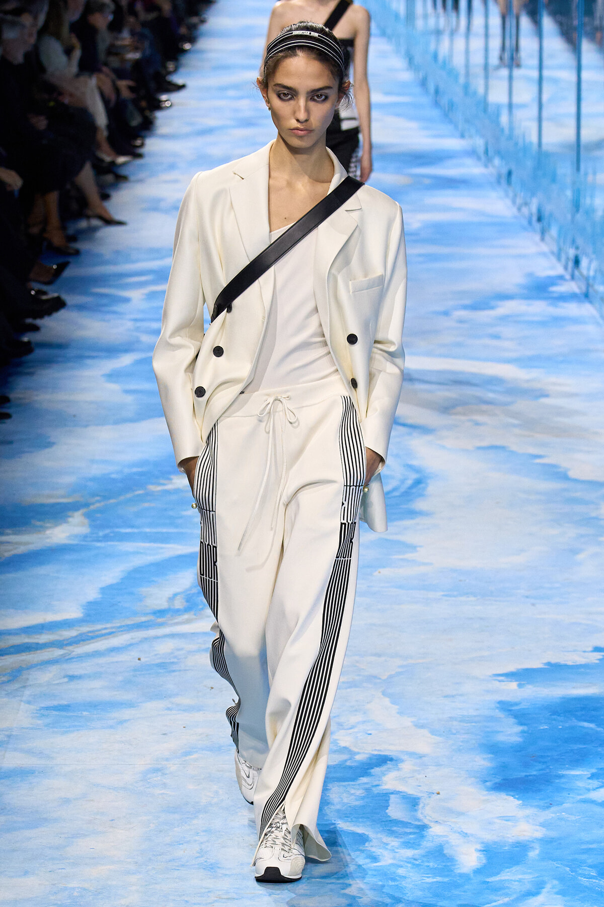 Model on a blue runway wearing a white blazer and drawstring white pants with black vertical stripes, and a black diagonal crossbody strap over the chest.