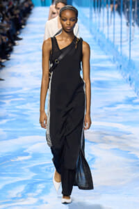 Female model walks a runway in a black sleeveless dress with white striped side panels and a crossbody bag.