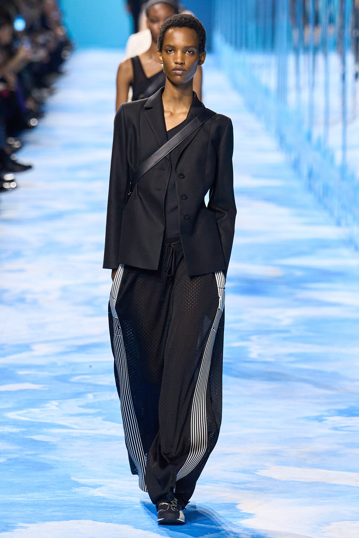 Model walking a blue-lit runway in a black blazer and loose black mesh pants with white striped sides.
