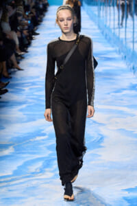 Model walks the blue runway in a black perforated jumpsuit with white-striped sleeves and a crossbody bag.