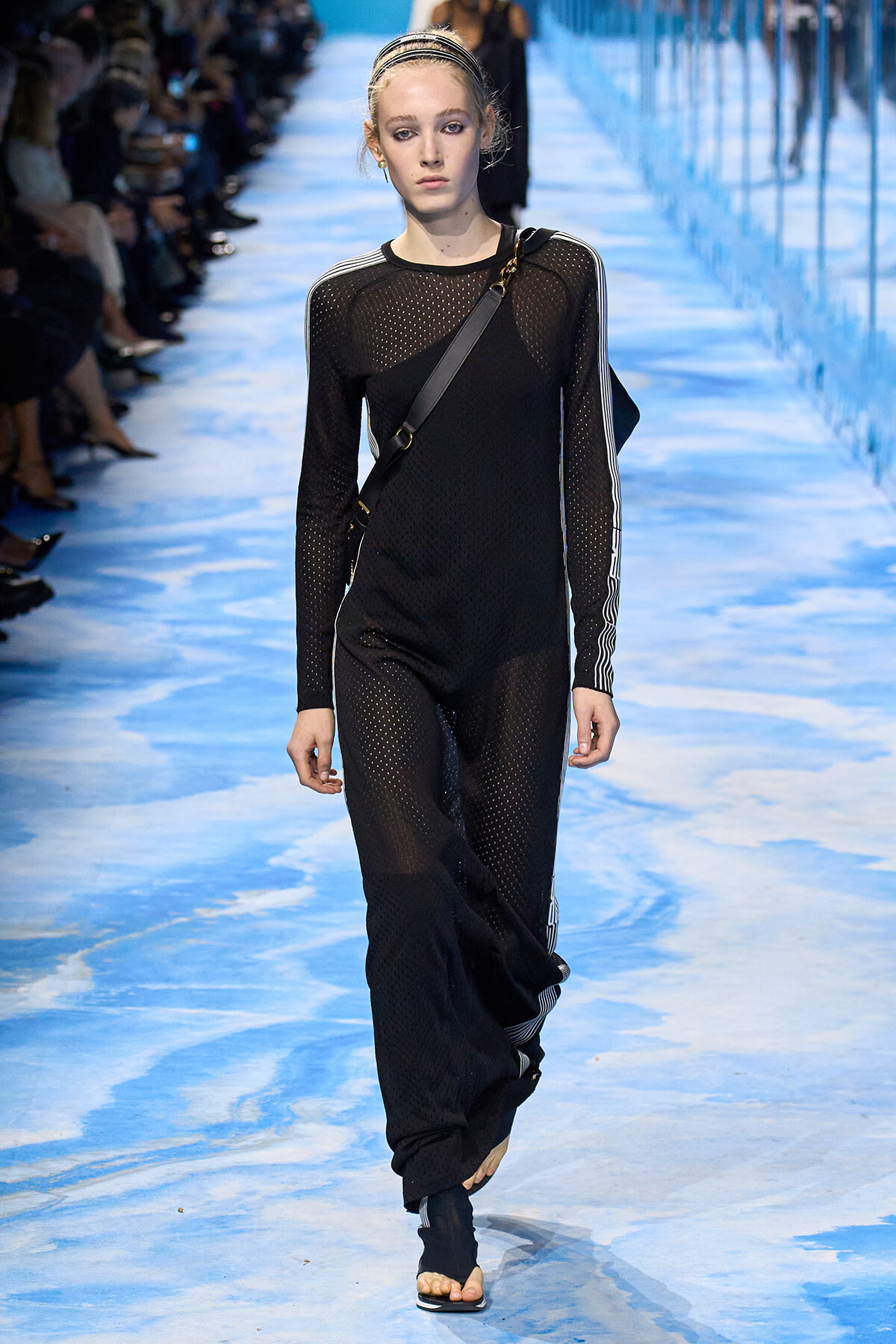 Model walks the blue runway in a black perforated jumpsuit with white-striped sleeves and a crossbody bag.