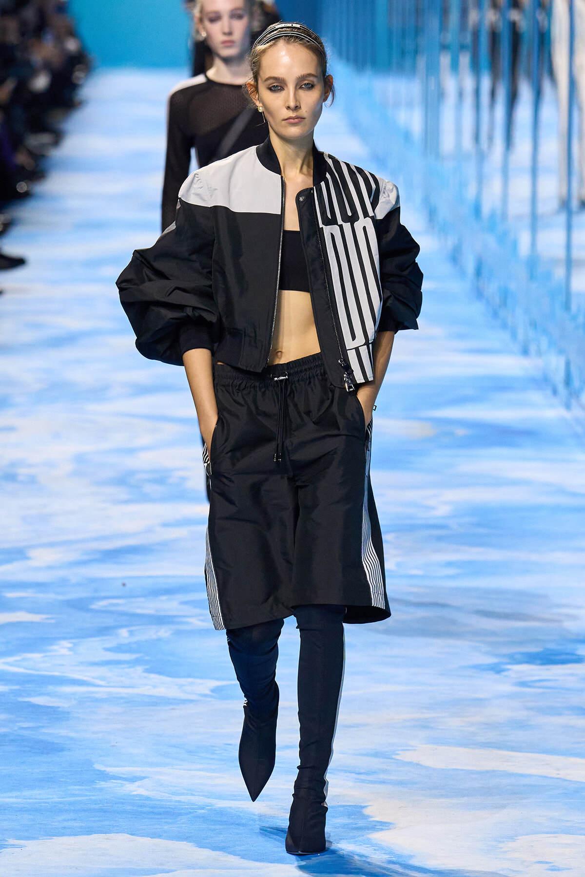 Model walking a blue-lit runway in a black and white color-block bomber jacket, black crop top, and black knee-length shorts with white side stripes, finished with black boots.