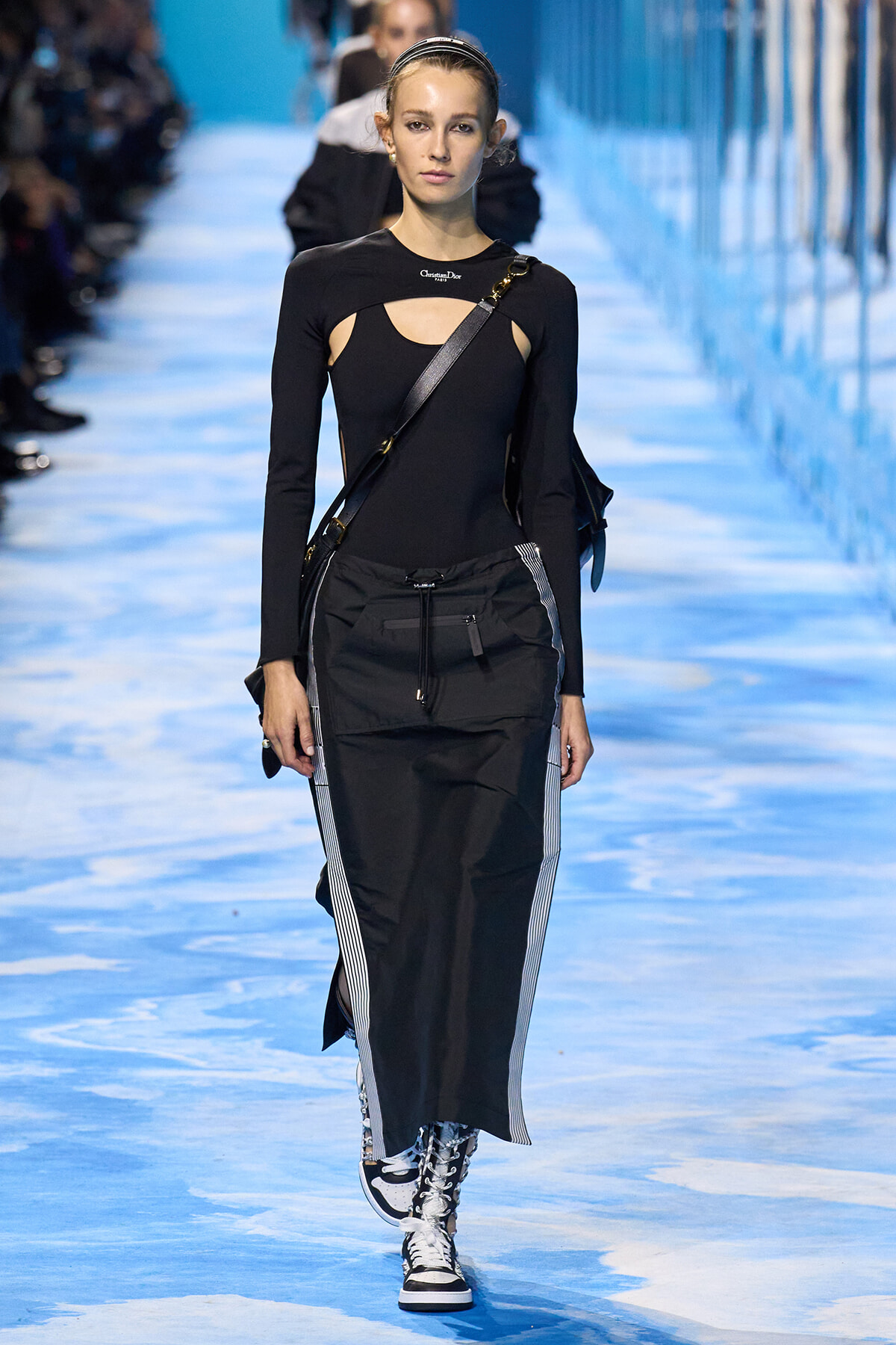 Model walking runway in black cutout top, black cargo skirt, crossbody bag, and white-black sneakers on blue-toned catwalk