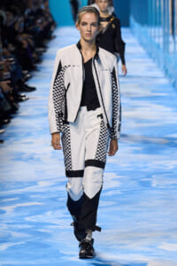 Model walks a white and black checkered uniform-style outfit on a blue runway, zippers visible.