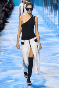 Model walks a blue runway in a black one-shoulder top and white split-front skirt with checkered trim, wearing oversized sunglasses.
