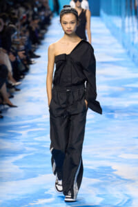 Female model walks a blue-toned runway in an all-black outfit: one-shoulder top with a draped sleeve and matching jogger pants, white sneakers, and a striped headband.