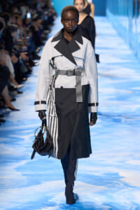 Model walking a fashion show in a black-and-white trench coat with a striped belt, holding a black handbag.
