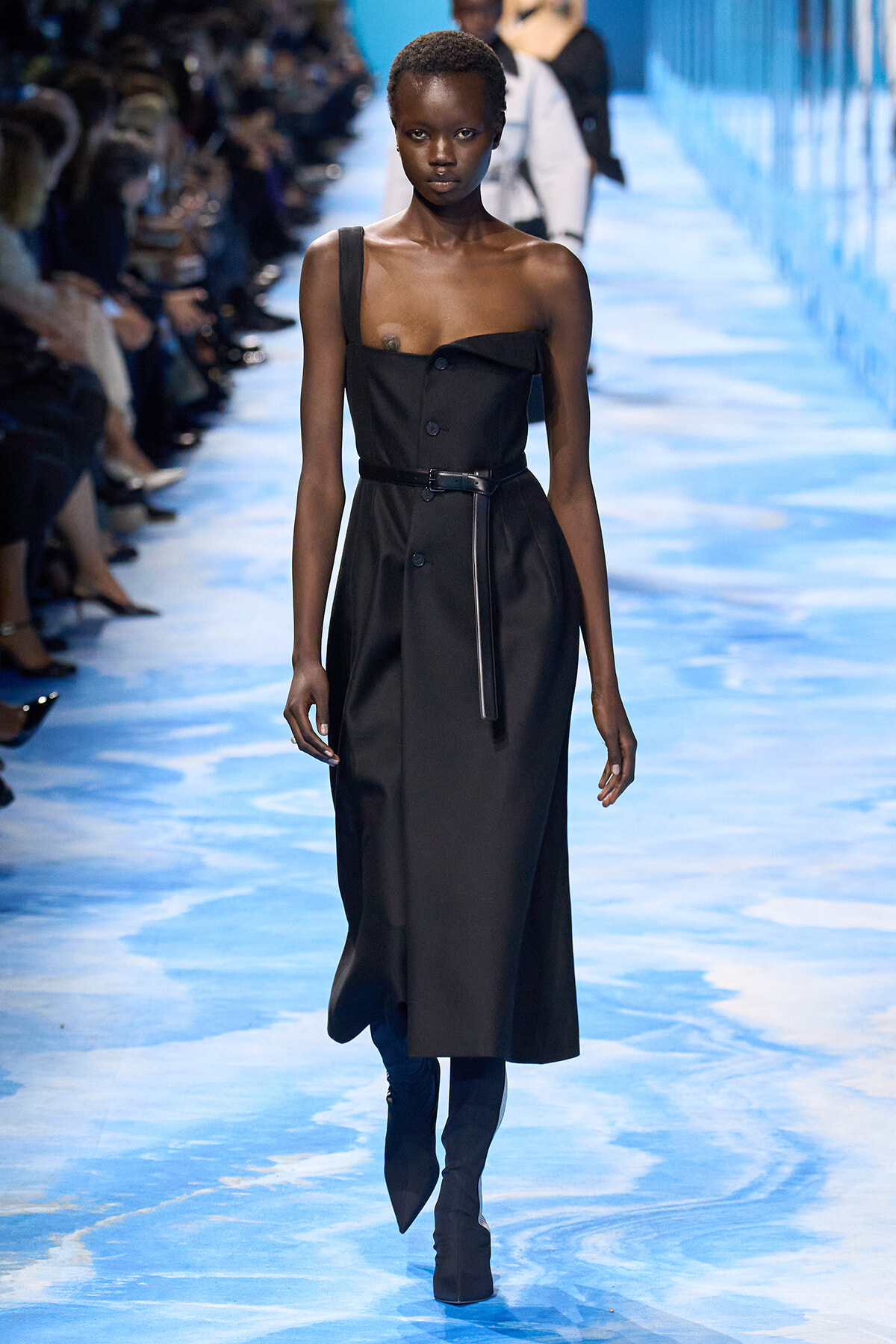 Black dress with a belt on a model walking a blue-lit runway, one-shoulder strap detail visible.