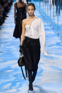 Runway model wearing a white one-shoulder blouse with black wide-leg pants, black gloves, and a small black handbag on a blue-toned runway.