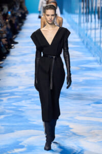 Model walks the runway in a black V-neck jumpsuit with a belt, long black gloves, and knee-high boots on a blue-toned catwalk.
