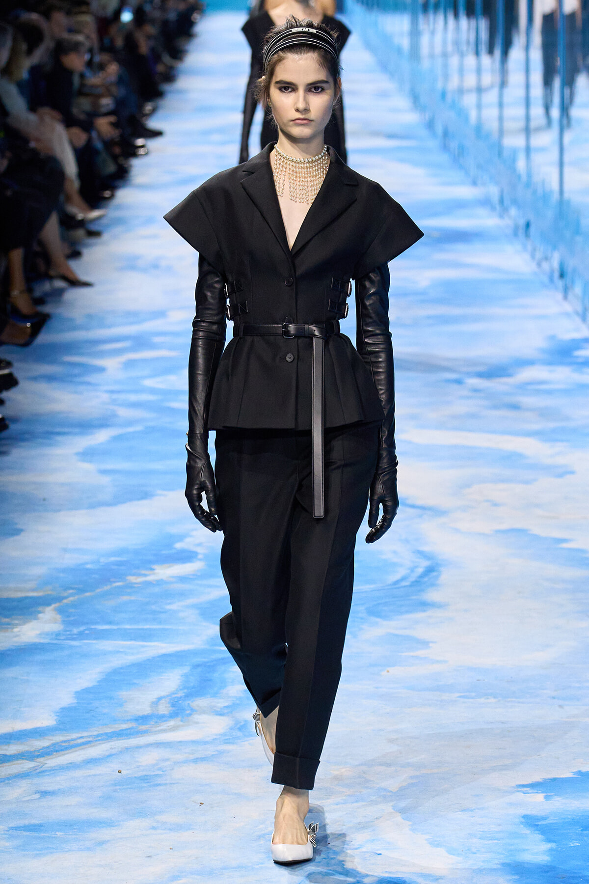 Model walks the runway in a black tailored ensemble with exaggerated shoulders, belt, and long leather gloves, accessorized with a pearl choker and headband.