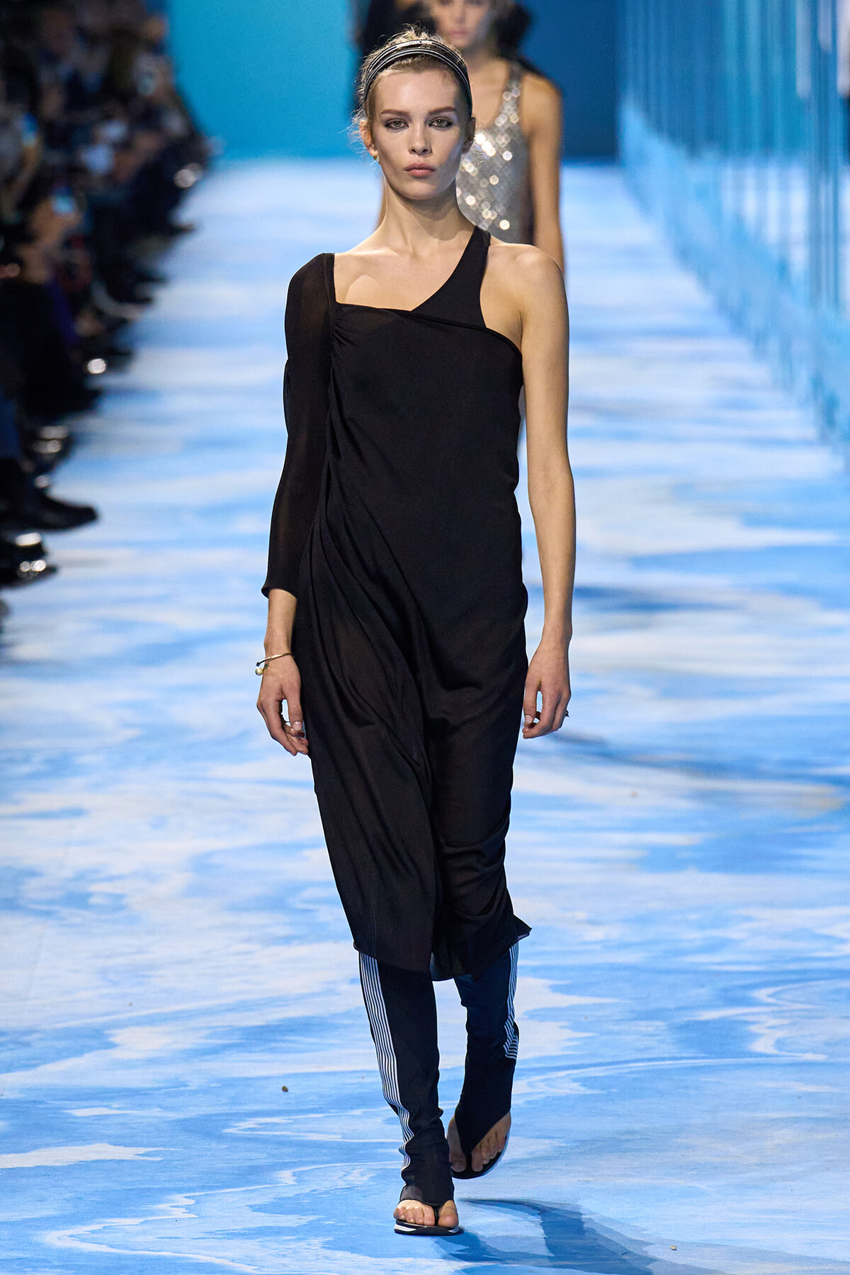 Model on a blue runway wearing a black one-shoulder draped dress with striped blue leggings and sandals.