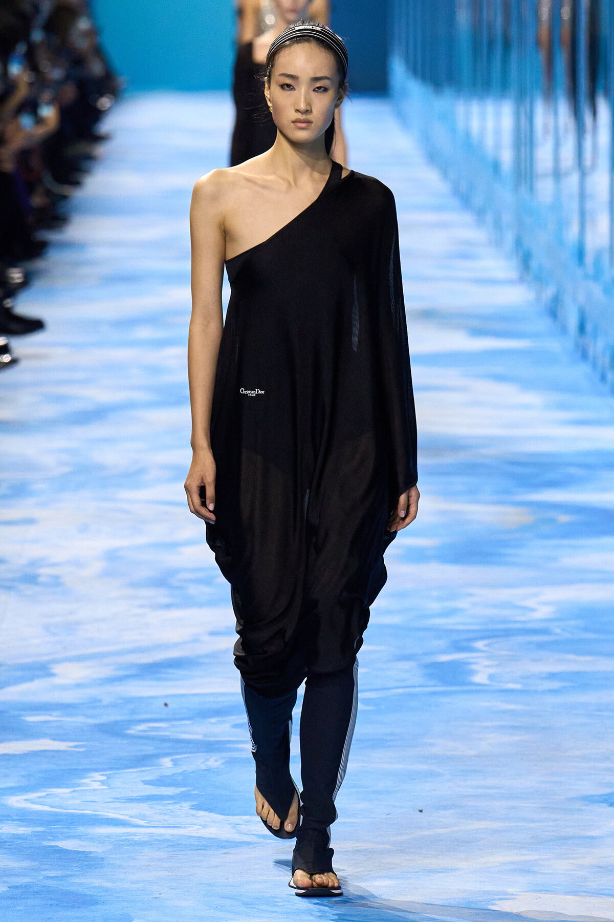 Model walking a fashion show in a black one-shoulder draped jumpsuit on a blue runway.