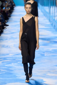 Model walks a fashion show wearing a black, draped, sleeveless dress with an asymmetric neckline on a blue runway.