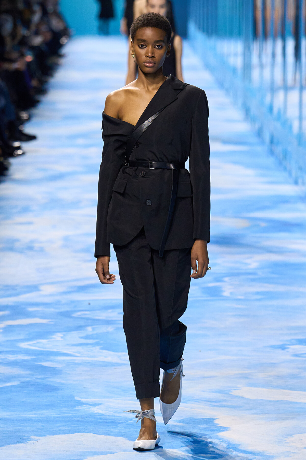 Female model walks the blue runway in a black one-shoulder blazer with a belt and matching trousers, white flats with ribbon ties.