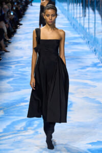 Female model walking a runway in a black one-shoulder midi dress with a side bow detail on a blue-lit catwalk