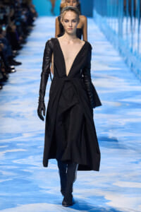 Model walks a black deep-V gown with long sleeves and gloves on a blue runway; headband completes look.