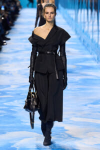 Model walks a black avant-garde suit with off-the-shoulder top, wide belt, and long gloves on a blue runway, carrying a black handbag.