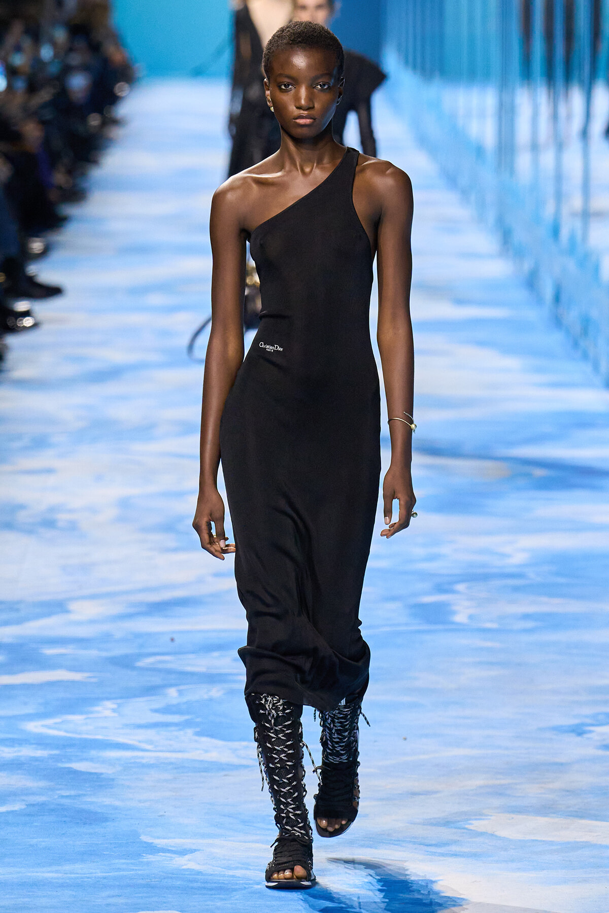 Model walking a blue-toned runway in a black one-shoulder dress and tall lace-up boots.