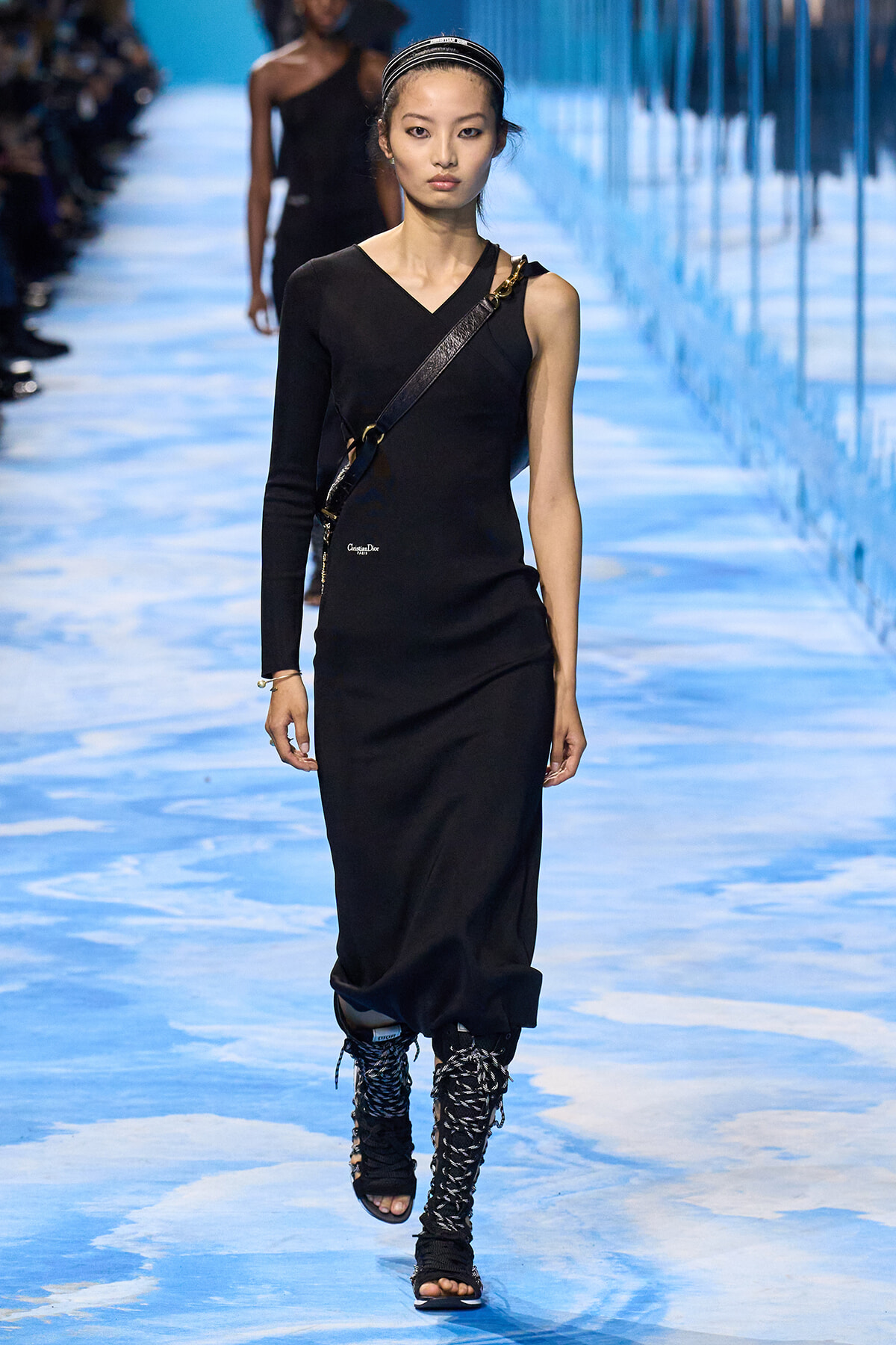 Model walking runway in a black one-shoulder dress with a crossbody strap, braided boots, and a striped headband on a blue-toned catwalk.