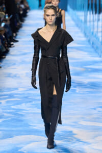 Model walking the runway in a black belted trench dress with long gloves and knee-high boots on a blue fashion catwalk.