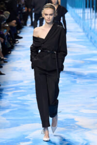 Model walking a runway in a black, one-shoulder blazer with a belt and matching trousers, white heels on a blue-toned catwalk.