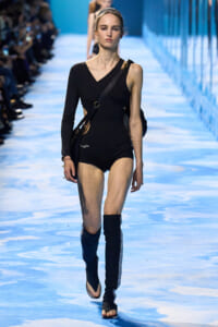 Female model walking a fashion show runway in a black asymmetrical one-piece with a single sleeve and thigh-high blue leg wraps.
