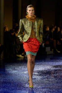 Runway model in a shimmering green jacket with an orange ruffle neck, a red ruched skirt with teal lace trim, and yellow heels on a glittery blue catwalk.
