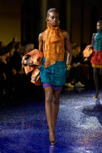 Model walking a fashion runway in an orange lace halter top and teal layered skirt, carrying a metallic orange coat.