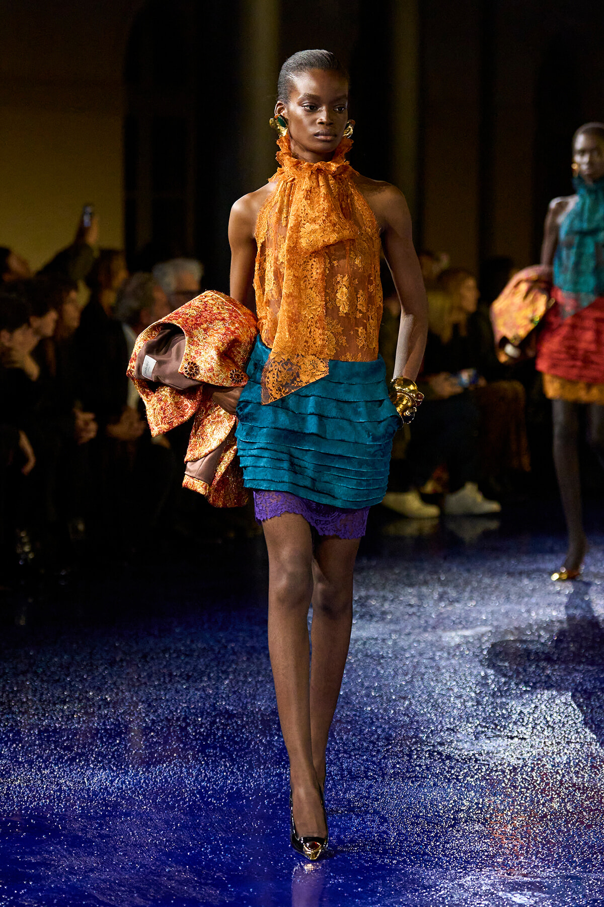 Model walking a fashion runway in an orange lace halter top and teal layered skirt, carrying a metallic orange coat.
