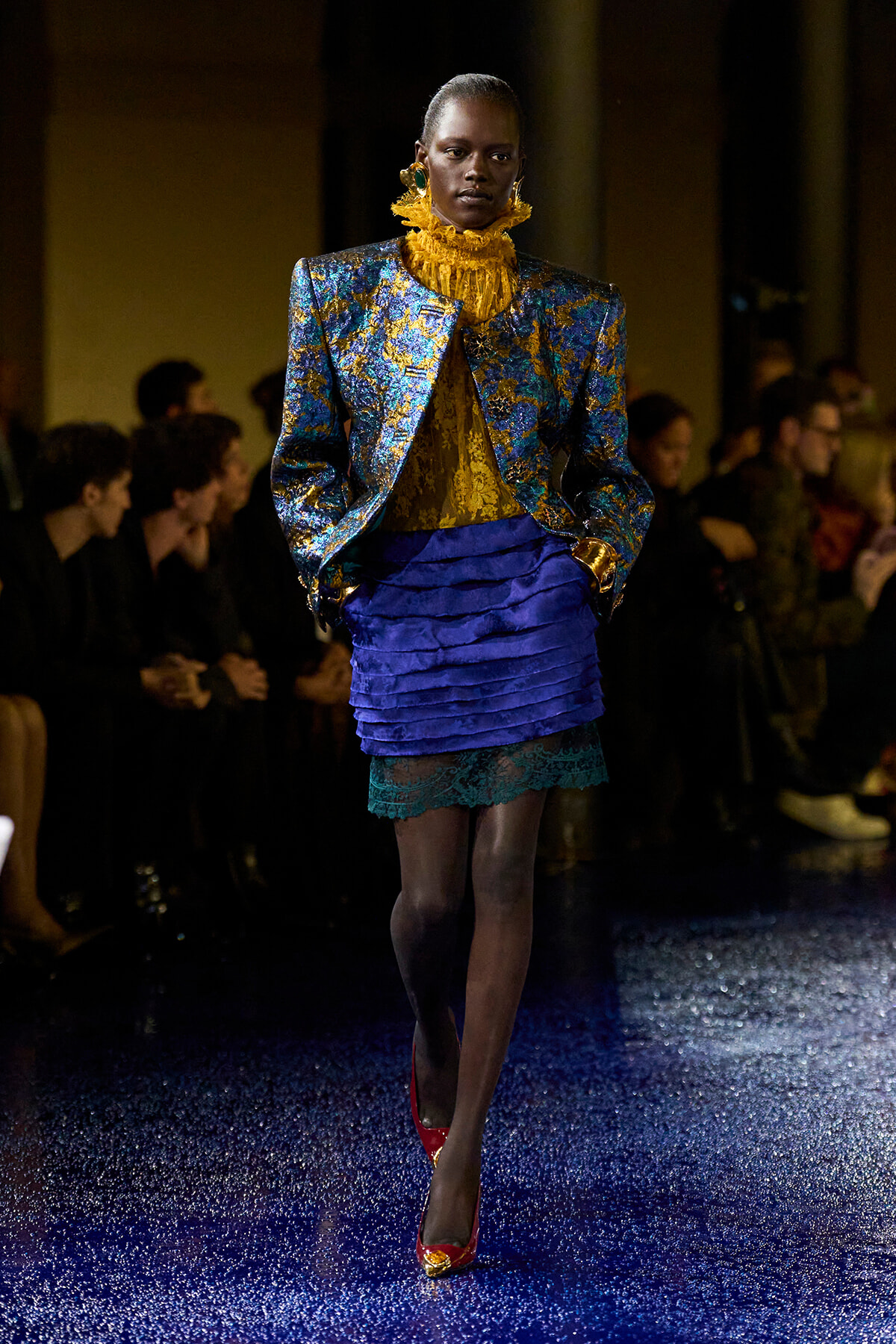 Model walking a fashion runway in a metallic blue-gold brocade blazer, yellow ruffled top, and blue tiered skirt with lace hem; red heels complement the look. Audience blurred in the background.