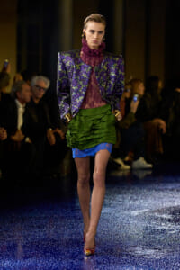 Model on a runway wearing a metallic purple-green jacket over a burgundy lace top, with a layered green skirt and blue lace hem.