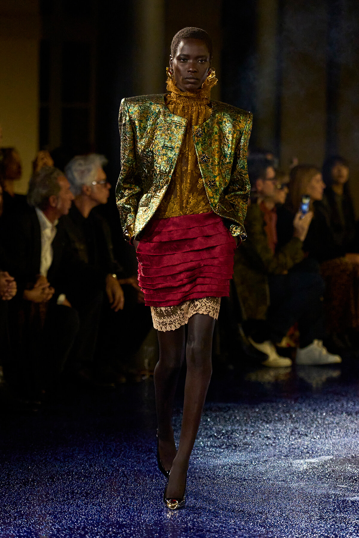 Female model walking a fashion show in a metallic gold-green blazer, gold high-neck top, and a red ruched mini skirt with lace trim; audience in background.