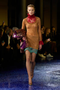 Model walking a runway in a shimmering peach-gold dress with red lace neck and teal lace hem, carrying a metallic jacket with purple lining, gold heels.