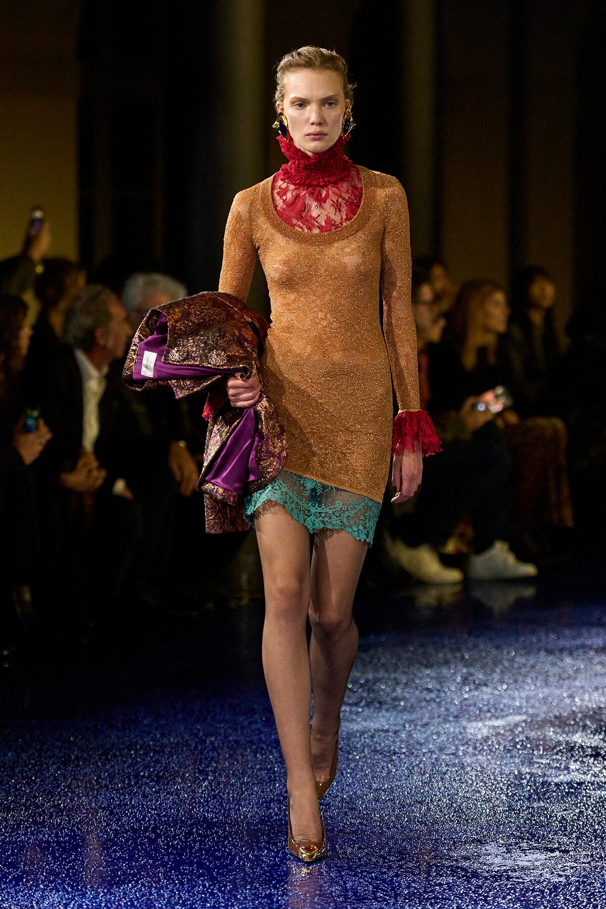 Model walking a runway in a shimmering peach-gold dress with red lace neck and teal lace hem, carrying a metallic jacket with purple lining, gold heels.