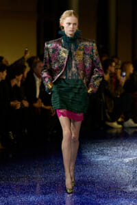 Model walks runway in a metallic floral blazer with teal lace blouse and a layered green skirt with pink lace at the hem, gold cuffs visible.