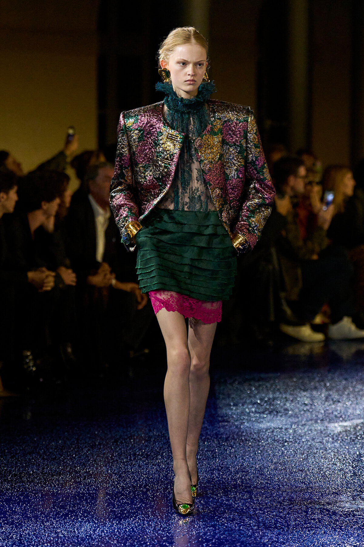Model walks runway in a metallic floral blazer with teal lace blouse and a layered green skirt with pink lace at the hem, gold cuffs visible.
