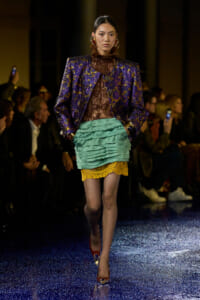 Model walking runway in a purple gold brocade blazer over a brown lace top, teal layered mini skirt with yellow lace trim, red-gold heels.