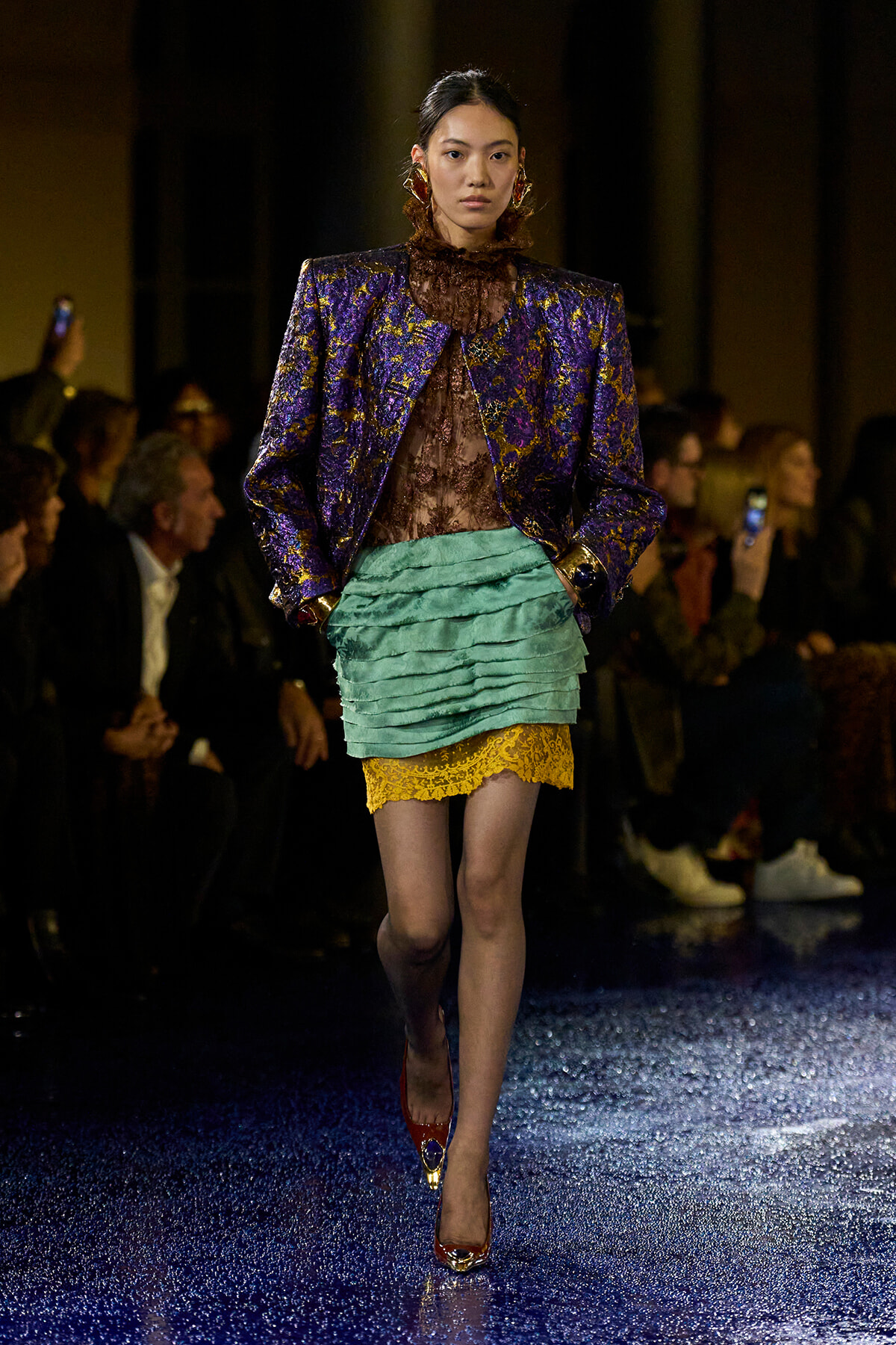 Model walking runway in a purple gold brocade blazer over a brown lace top, teal layered mini skirt with yellow lace trim, red-gold heels.