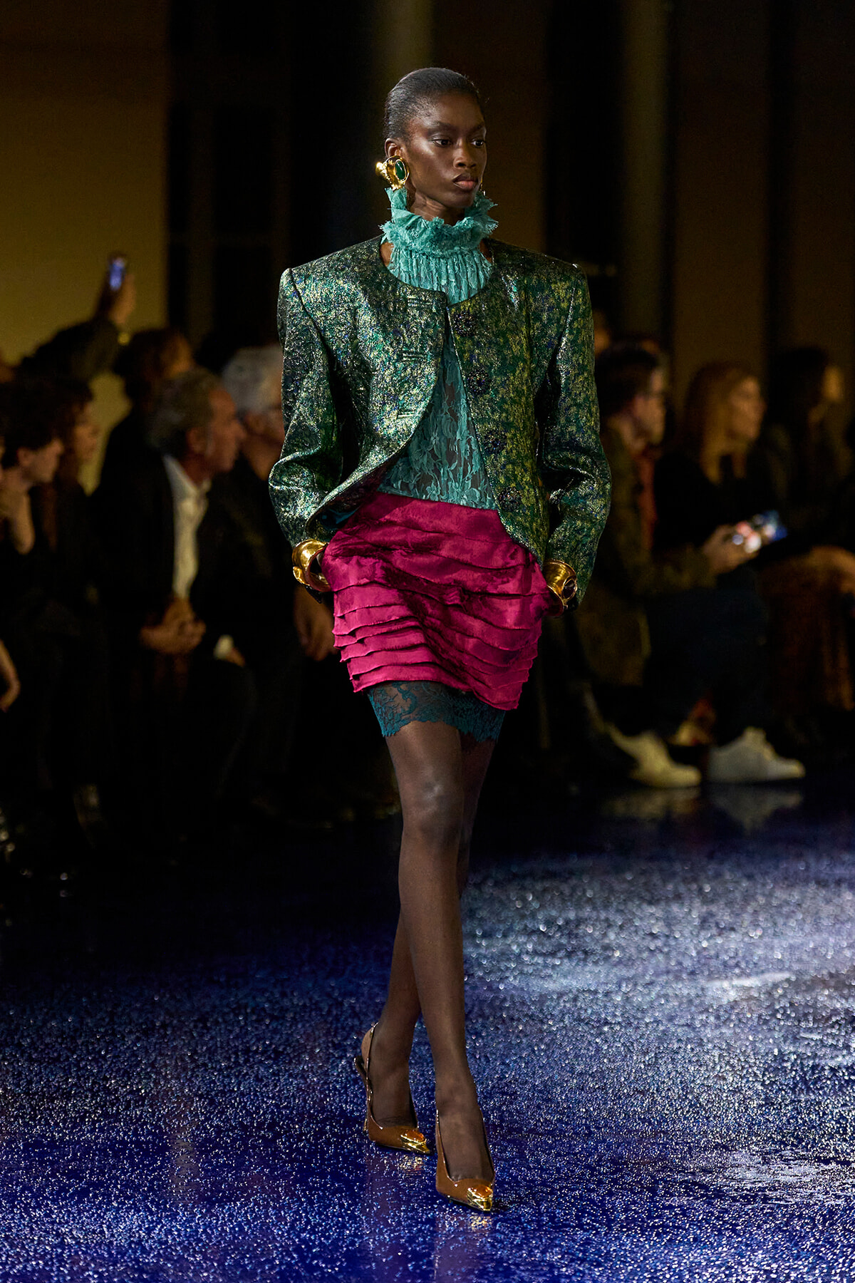 Model walking a fashion show in a green metallic blazer, teal ruffled high-neck blouse, and magenta layered mini skirt with teal lace tights and gold heels.
