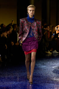 Model walks a glittery blue runway in a metallic burgundy-green patterned blazer, royal blue lace collar top, and a layered purple mini with red lace trim.