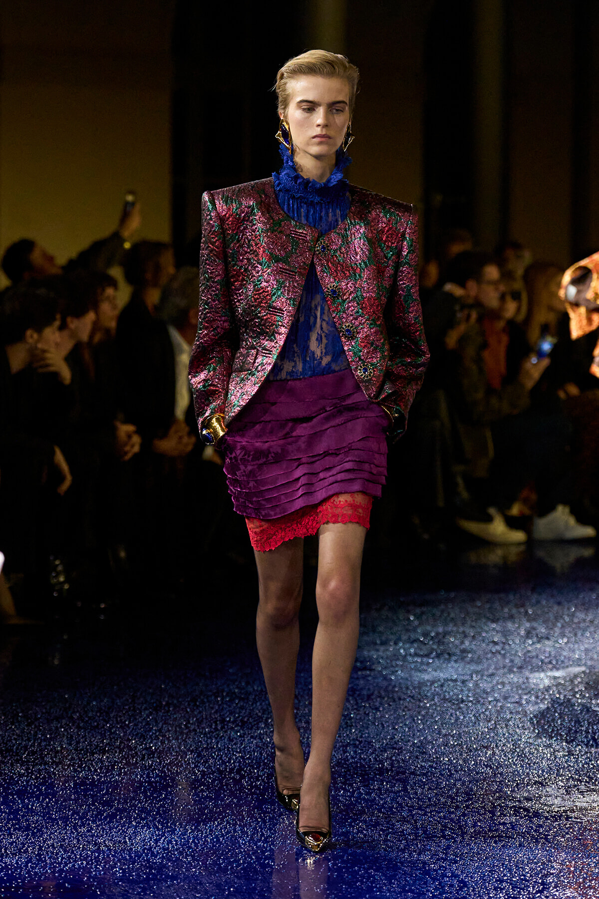 Model walks a glittery blue runway in a metallic burgundy-green patterned blazer, royal blue lace collar top, and a layered purple mini with red lace trim.