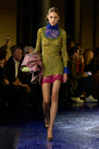 Model walking a runway in a glittery olive-gold mini dress with a blue lace high neck, pink lace hem, carrying a metallic pink-green jacket.