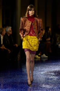 Model walking a fashion runway in a metallic burgundy blazer over a red lace blouse, paired with a bright yellow tiered skirt and purple lace shorts, black high heels, audience in the background.