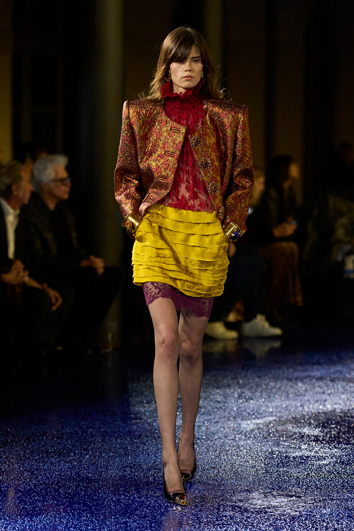Model walking a fashion runway in a metallic burgundy blazer over a red lace blouse, paired with a bright yellow tiered skirt and purple lace shorts, black high heels, audience in the background.