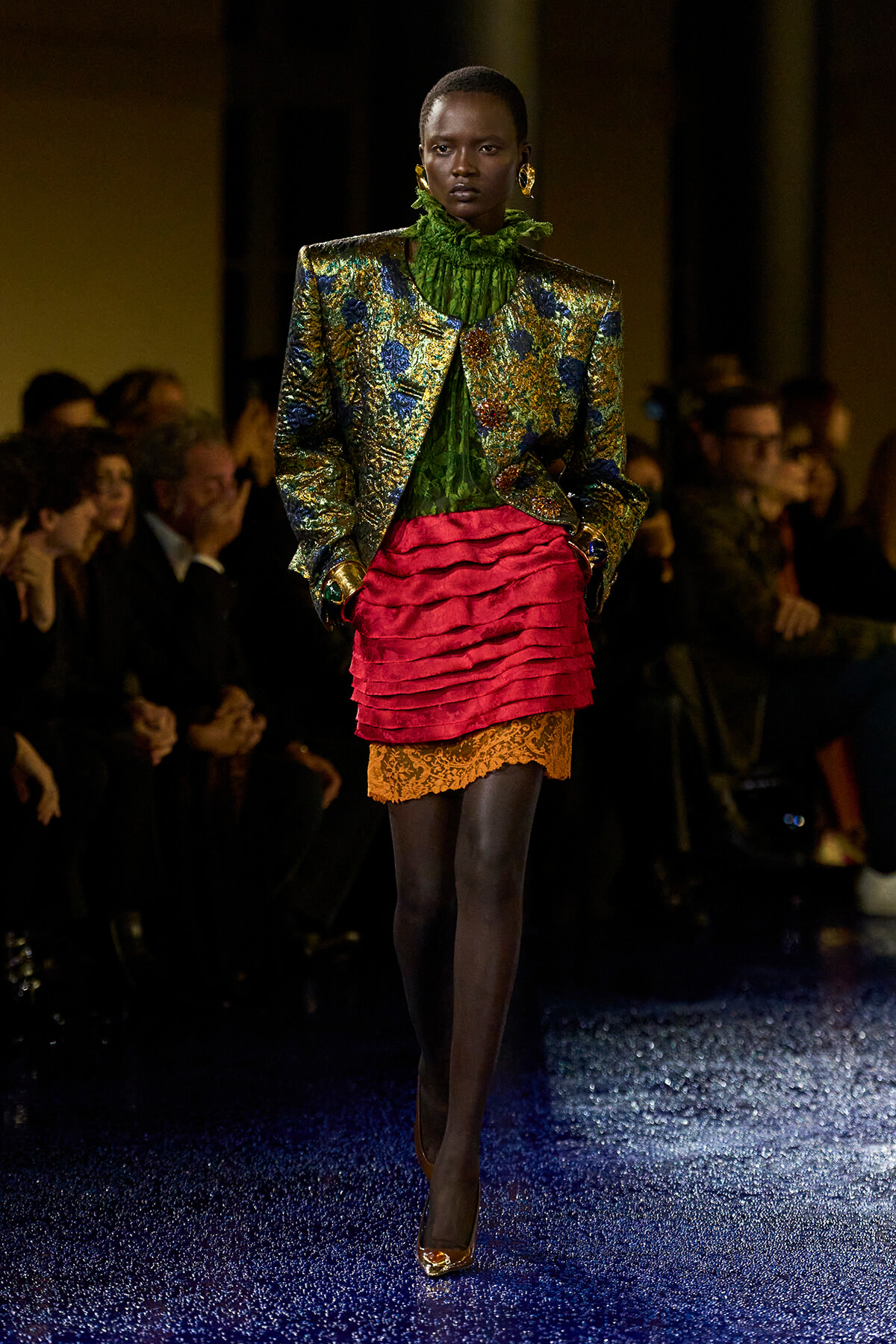 Model walks the runway in a metallic floral brocade jacket over a green ruffled blouse and a pink layered mini skirt with orange lace trim.