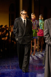 Model walks a runway in an oversized black double-breasted suit, white shirt, and black tie, wearing glasses against a glittery blue floor.