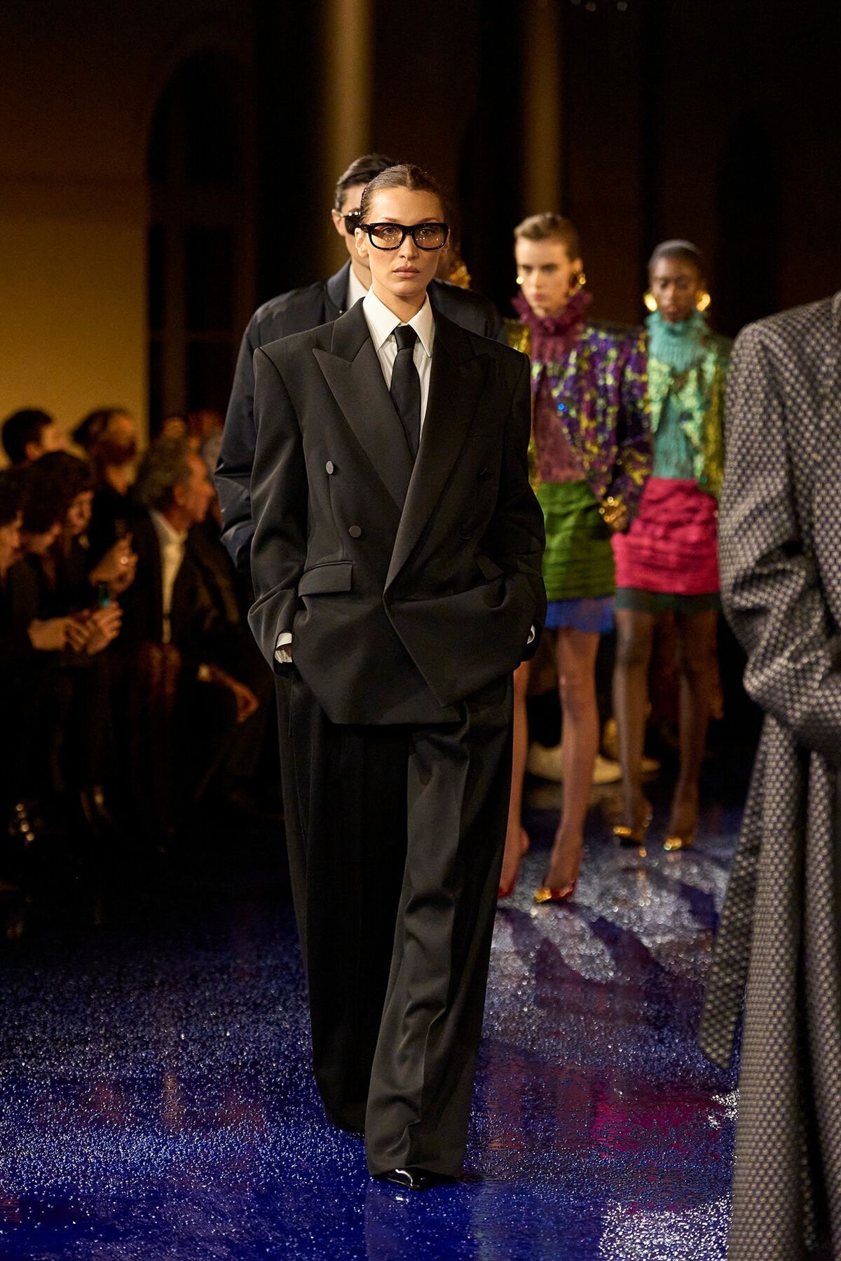 Model walks a runway in an oversized black double-breasted suit, white shirt, and black tie, wearing glasses against a glittery blue floor.