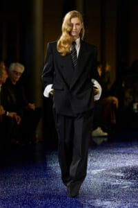 Female model walks a fashion runway in an oversized black double-breasted suit with white cuffs and a striped tie on a glittery blue catwalk.